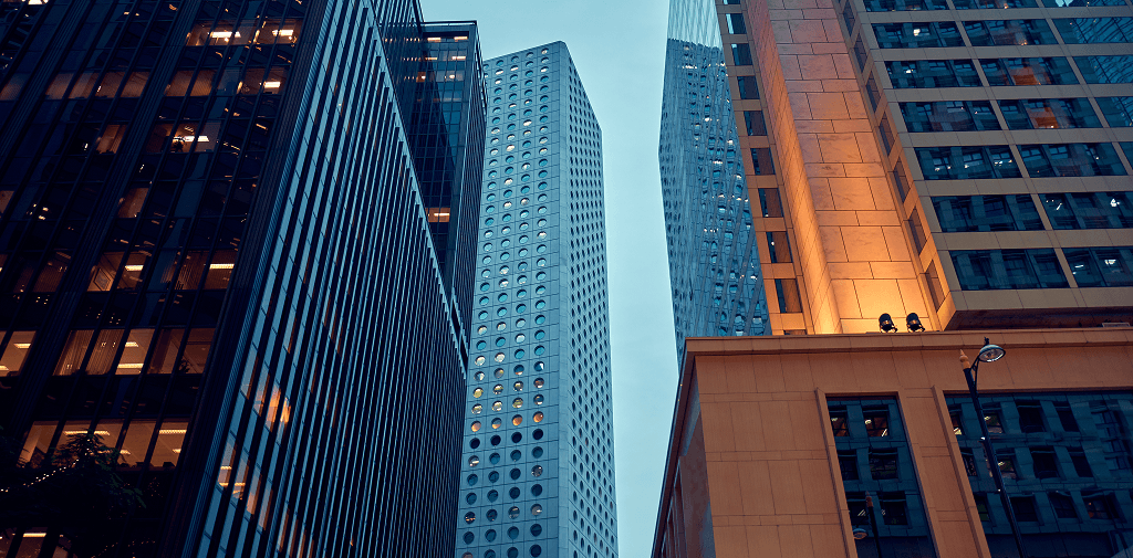 Une vue en contre-plongée de gratte-ciel urbains modernes, mettant en valeur une tour blanche centrale aux fenêtres circulaires distinctives, entourée d'immeubles de bureaux en verre sombre et chaleureusement éclairés sous un ciel de crépuscule.